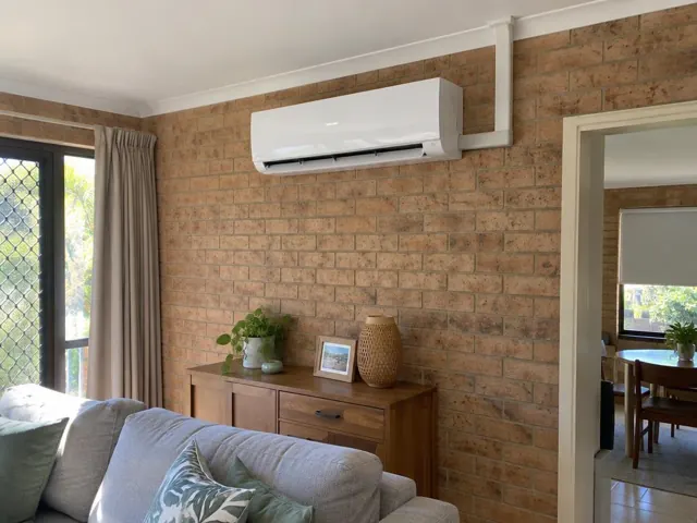 Wall-mounted split system above a brick feature wall in a living room with sofa and cabinet in Coomera